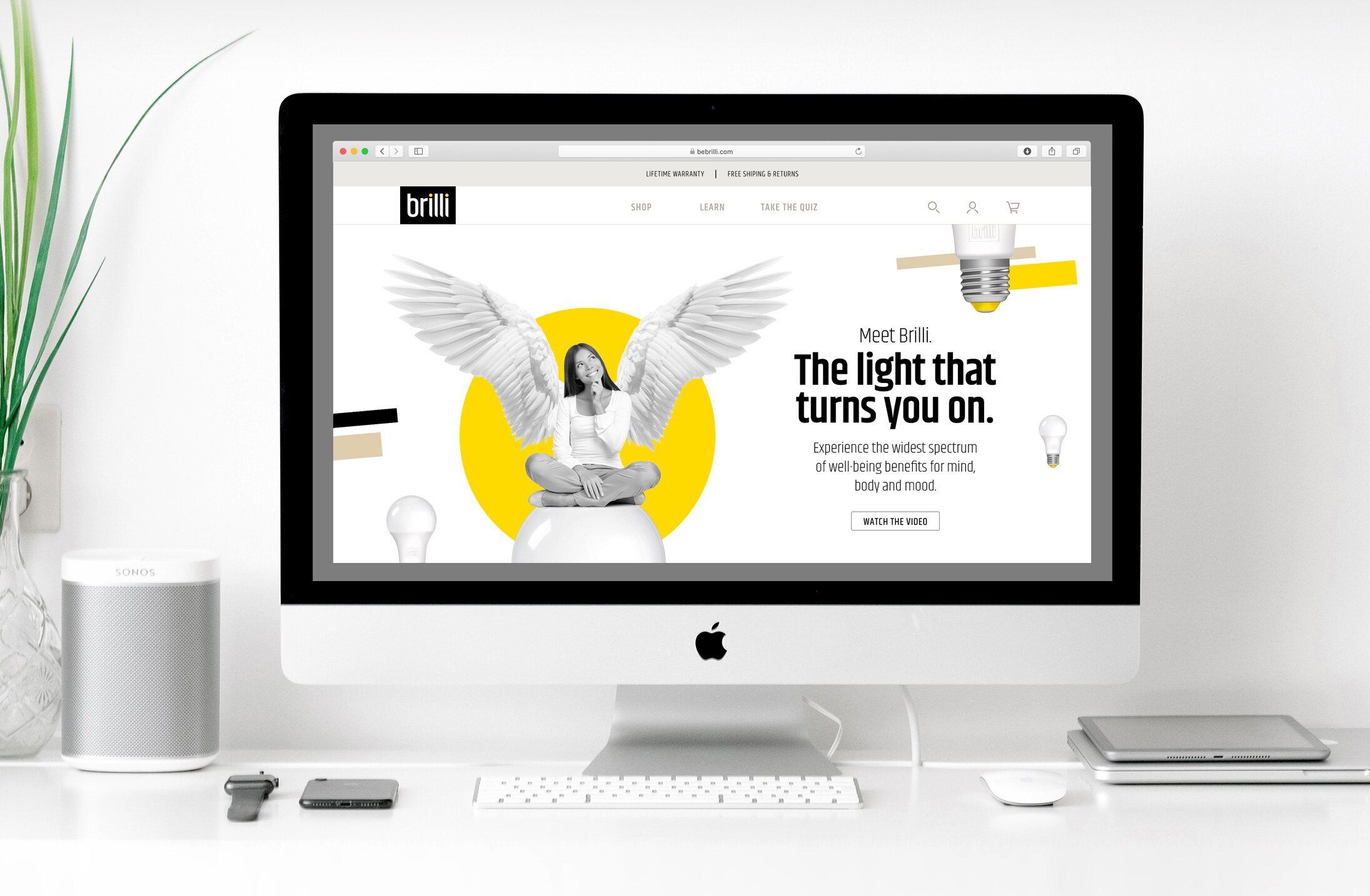 An Apple iMac on a white desk displaying a website for a lighting company called Brilli, featuring a woman with angel wings sitting cross-legged, with text promoting the benefits of their lights. There is a Sonos speaker, an Apple Watch, a smartphone